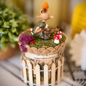 May include: A cylindrical decorative container featuring a miniature figurine of a creature with orange hair, a green top, and a small bucket. The top is covered in green moss, purple lichen, and red-and-white mushrooms, surrounded by a wooden picket fence.