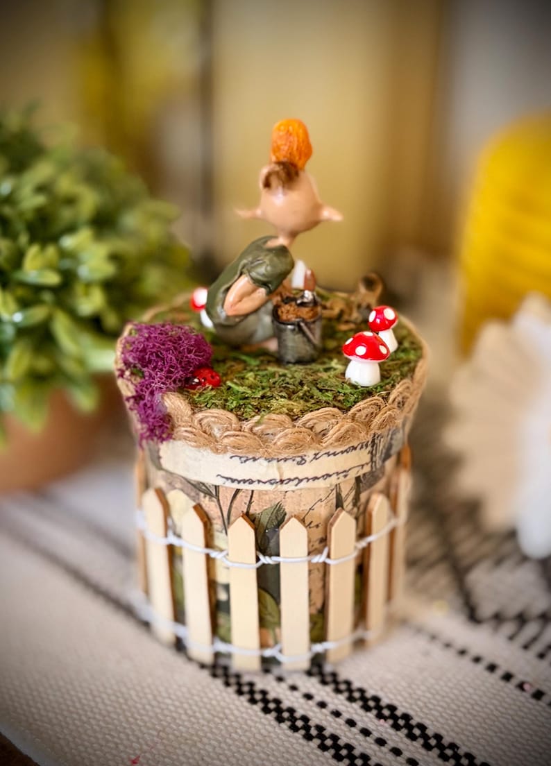 May include: A cylindrical decorative container featuring a miniature figurine of a creature with orange hair, a green top, and a small bucket. The top is covered in green moss, purple lichen, and red-and-white mushrooms, surrounded by a wooden picket fence.