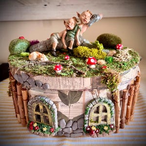 May include: A decorative round box featuring a miniature scene on top with two small elf-like figures resting on a log, surrounded by green moss, tiny red and white mushrooms, and a small animal. The sides are decorated with paper, twig fencing, and two arched windows with flower boxes.