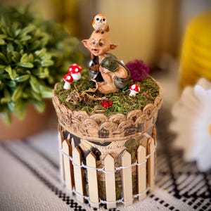 May include: A cylindrical decorative container featuring a miniature elf figure with an owl on its head, surrounded by small red and white mushrooms on a mossy top. The base is wrapped in a miniature wooden picket fence with white wire accents.