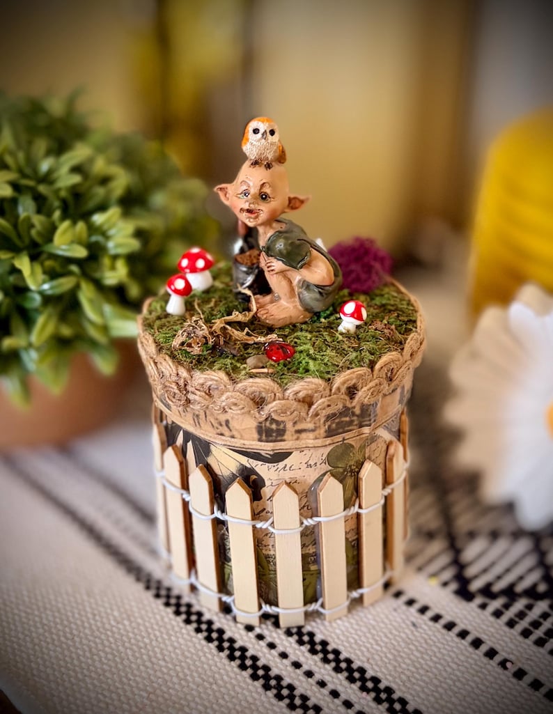 May include: A cylindrical decorative container featuring a miniature elf figure with an owl on its head, surrounded by small red and white mushrooms on a mossy top. The base is wrapped in a miniature wooden picket fence with white wire accents.