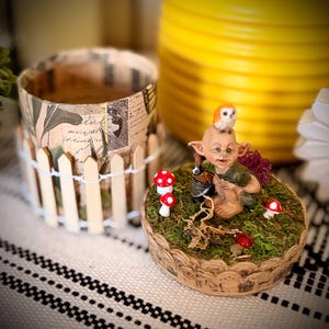 May include: A decorative set featuring a cylindrical container wrapped in vintage-style paper with a miniature picket fence, and a circular base covered in green moss, small red mushrooms, a brown creature figurine, and a tiny owl on its head.
