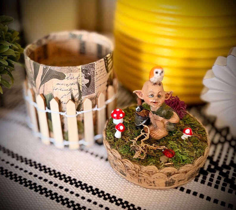 May include: A decorative set featuring a cylindrical container wrapped in vintage-style paper with a miniature picket fence, and a circular base covered in green moss, small red mushrooms, a brown creature figurine, and a tiny owl on its head.