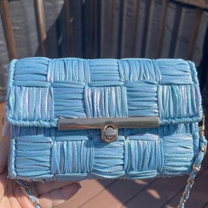 May include: A light blue rectangular handbag featuring a woven, ruched fabric texture with a subtle iridescent sheen. The bag includes a gold-toned metal bar clasp closure on the front flap and a matching light blue and gold-toned chain shoulder strap.