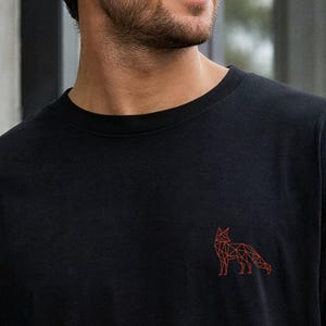 Könnte beinhalten: Ein schwarzes Kurzarm-T-Shirt mit einem kleinen, geometrischen Fuchsmotiv, das in einem feinen, rostorangen Linienstil auf der linken Brustseite dargestellt ist. Das Kleidungsstück verfügt über einen klassischen Rundhalsausschnitt und eine lockere Passform.