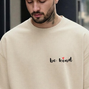 Pode incluir: Uma t-shirt de manga curta bege com o texto "be kind" em letra cursiva preta na parte superior esquerda do peito, com um pequeno ícone de coração vermelho posicionado acima da letra i. A t-shirt tem gola redonda e um corte largo.