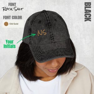 Op de afbeelding: Een zwarte acid-wash baseballpet met de geborduurde initialen NS in oudgoud draad. De tekst Rock Salt geeft de letterstijl aan en de tekst Your Initials wijst met een groene pijl naar het borduurwerk.