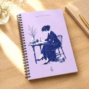 May include: A spiral-bound notebook with a lavender cover featuring an illustration of a person writing at a table. The text "she keeps notes." is printed at the top. A pen and a closed notebook are visible.