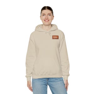 May include: A beige hooded sweatshirt with a front pocket and a drawstring hood. A small rectangular patch with text is on the upper chest. The model is wearing blue jeans. The sweatshirt is a casual garment.