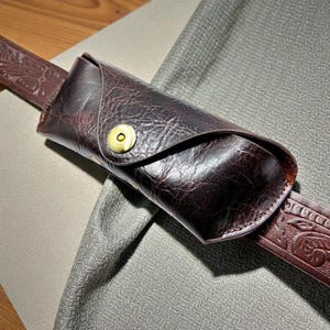 May include: A dark brown leather glasses case with a snap closure, attached to a matching embossed leather belt. The case has a textured surface and a gold-coloured snap. The belt is also dark brown.