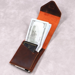 May include: A brown leather wallet with an orange interior, open to reveal a stack of $100 bills and a card. The wallet has a simple, minimalist design and appears to be handmade.