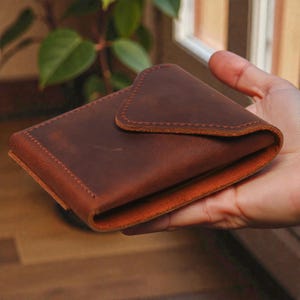 May include: A brown leather wallet with a folded flap closure. The wallet has a distressed finish and visible stitching around the edges. The interior appears to be a lighter shade of brown. The wallet is held in a hand.