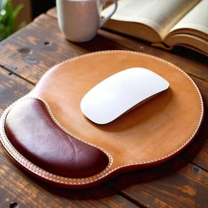 Pode incluir: Um mouse pad de couro castanho com um apoio de pulso bordô e um mouse sem fio branco. A almofada tem uma borda costurada. Uma caneca branca e um livro aberto estão no fundo. Sobre uma superfície de madeira.