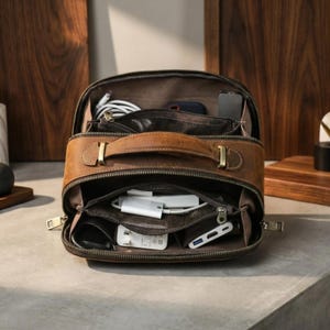 May include: Open brown leather travel organiser with multiple compartments, zips, and a top handle. The organiser is filled with various electronic accessories, including cables, chargers, and a power adapter. The bag is open, revealing its contents.