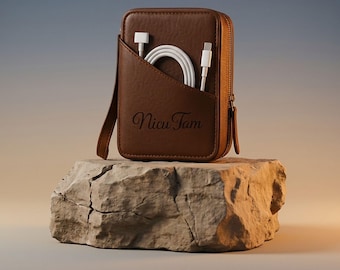 May include: A brown leather cable organizer with a zipper closure and a front pocket. The organizer contains a white charging cable. The text "Nicu Tam" is printed on the front. The organizer is displayed on a stone.