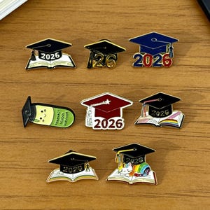 May include: A collection of graduation-themed enamel pins. Designs include graduation caps with the year 2026, open books, and a pharmacy school survivor pin. The pins feature a variety of colors, including black, blue, red, and white.