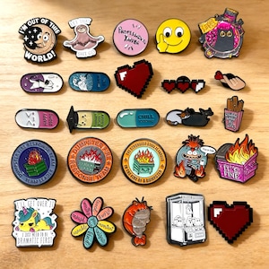 May include: A collection of enamel pins with various designs and text. The pins feature a pixelated heart, a smiling face, a sloth, a unicorn, and phrases like "I'm out of this world!" and "It's fine."