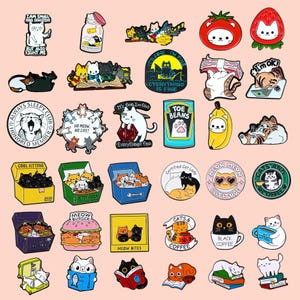 Könnte beinhalten: Eine Sammlung farbenfroher Emaille-Anstecknadeln zum Thema Katze. Designs zeigen Katzen in Kisten, beim Lesen von Büchern und in Lebensmitteln wie einem Burger und einer Banane. Einige Anstecknadeln haben Text wie "Everything is Fine".