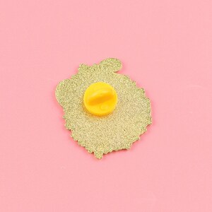 May include: A gold-colored enamel pin with a glittery texture. The pin features a yellow clutch back and a detailed design. The pin is set against a pink background, highlighting its color and design.