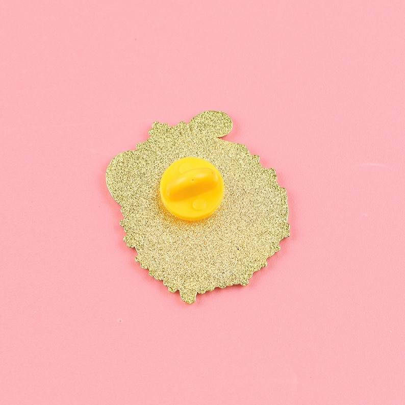 May include: A gold-colored enamel pin with a glittery texture. The pin features a yellow clutch back and a detailed design. The pin is set against a pink background, highlighting its color and design.