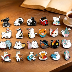 May include: A selection of cat-themed enamel pins in various colours. Each pin showcases a different cat design, such as cats with books, musical instruments, and coffee cups. The pins are displayed on a wooden surface, with a book and a cup of coffee visible.