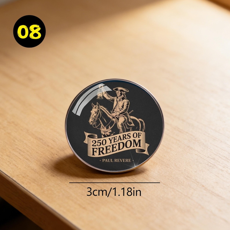 May include: A round, black button with a gold and white illustration of Paul Revere on horseback, holding a lantern. The text reads "250 YEARS OF FREEDOM - PAUL REVERE." The button is 1.18 inches in diameter.