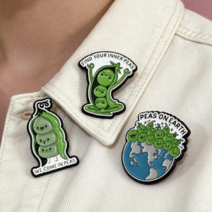 以下が含まれることがあります： エナメルピン3個セット。エンドウ豆のデザインで、「Find your inner peas」、「We come in peas」、「Peas on Earth」と書かれています。ピンには、笑顔の緑色のエンドウ豆と黒いアウトラインが描かれています。