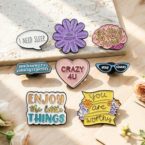 Peut inclure: Collection d'épingles émaillées avec des phrases et des motifs motivants. Comprend une bulle de dialogue avec "I NEED SLEEP", une fleur avec des mots comme "love" et "patience", et un cerveau avec "BE KIND TO YOUR MIND".