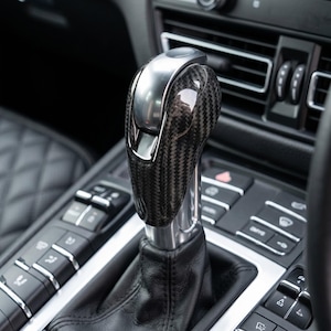 May include: A car gear stick with a carbon fibre and chrome finish. The surrounding interior features black leather and silver buttons. The gear stick is positioned in the centre console of the car.