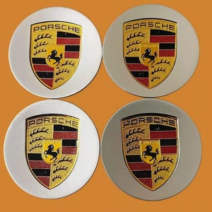 May include: Four silver-colored Porsche wheel center caps. Each cap features the Porsche emblem: a black horse on a yellow background, with red and black stripes, and the word "Porsche" in black.