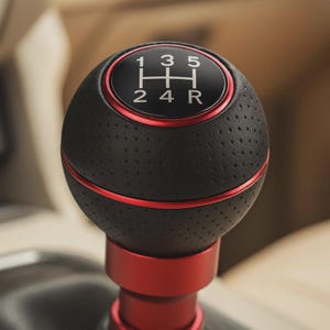 May include: A black and red car gear shift knob. The top is black with white numbers and a gear pattern. The knob has a perforated texture and a red accent ring. The base is red.