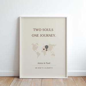 Könnte beinhalten: Gerahmter Kunstdruck mit dem Text „TWO SOULS ONE JOURNEY.“ Eine Weltkarten-Grafik zeigt einen Heißluftballon und ein Herz. Die Namen „Anna & Paul“ und Koordinaten stehen darunter. Der Druck ist in einem weißen Rahmen.