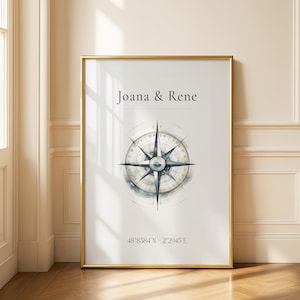 Könnte beinhalten: Gerahmter Druck mit einem Kompassrosen-Design in Blau- und Grautönen, mit den Namen "Joana & Rene" oben. Koordinaten sind unten aufgedruckt. Der Rahmen ist goldfarben.