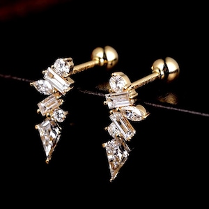 May include: A pair of gold-colored stud earrings with a cluster of clear, sparkling crystals. The crystals are various shapes, including round, baguette, and teardrop, arranged in a delicate, asymmetrical design. The earrings have a post and ball backing.