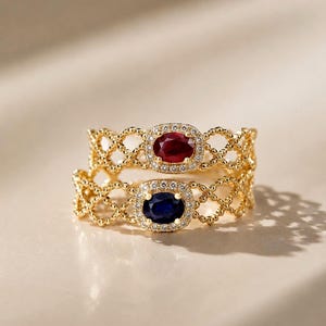 May include: Two gold rings featuring an intricate lattice band design. The top ring holds an oval-cut red gemstone, while the bottom ring features an oval-cut blue gemstone. Both center stones are surrounded by a halo of small, clear white stones.