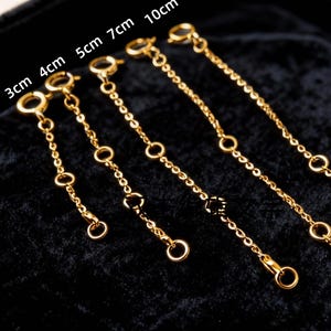 May include: Gold-colored necklace extenders in 3cm, 4cm, 5cm, 7cm, and 10cm lengths. Each extender features a small clasp on one end and a ring on the other, with additional rings along the chain. The extenders are displayed on a dark velvet surface.
