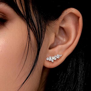May include: A close-up of a person's ear adorned with a delicate, asymmetrical earring. The earring features multiple clear, sparkling gemstones in various shapes, including round and baguette cuts, arranged in a cluster design.