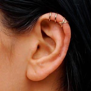 May include: Close-up of an ear adorned with multiple gold-colored earrings. Three small hoops are stacked on the upper ear, and a criss-cross design hoop is also visible. A single gold stud earring is in the earlobe.