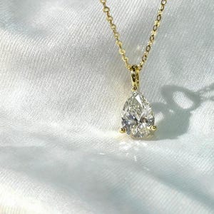 May include: A gold necklace with a pear-shaped diamond pendant. The diamond is clear and faceted, reflecting light. The chain is delicate and gold, with a simple clasp. The necklace is displayed on a white, textured surface.