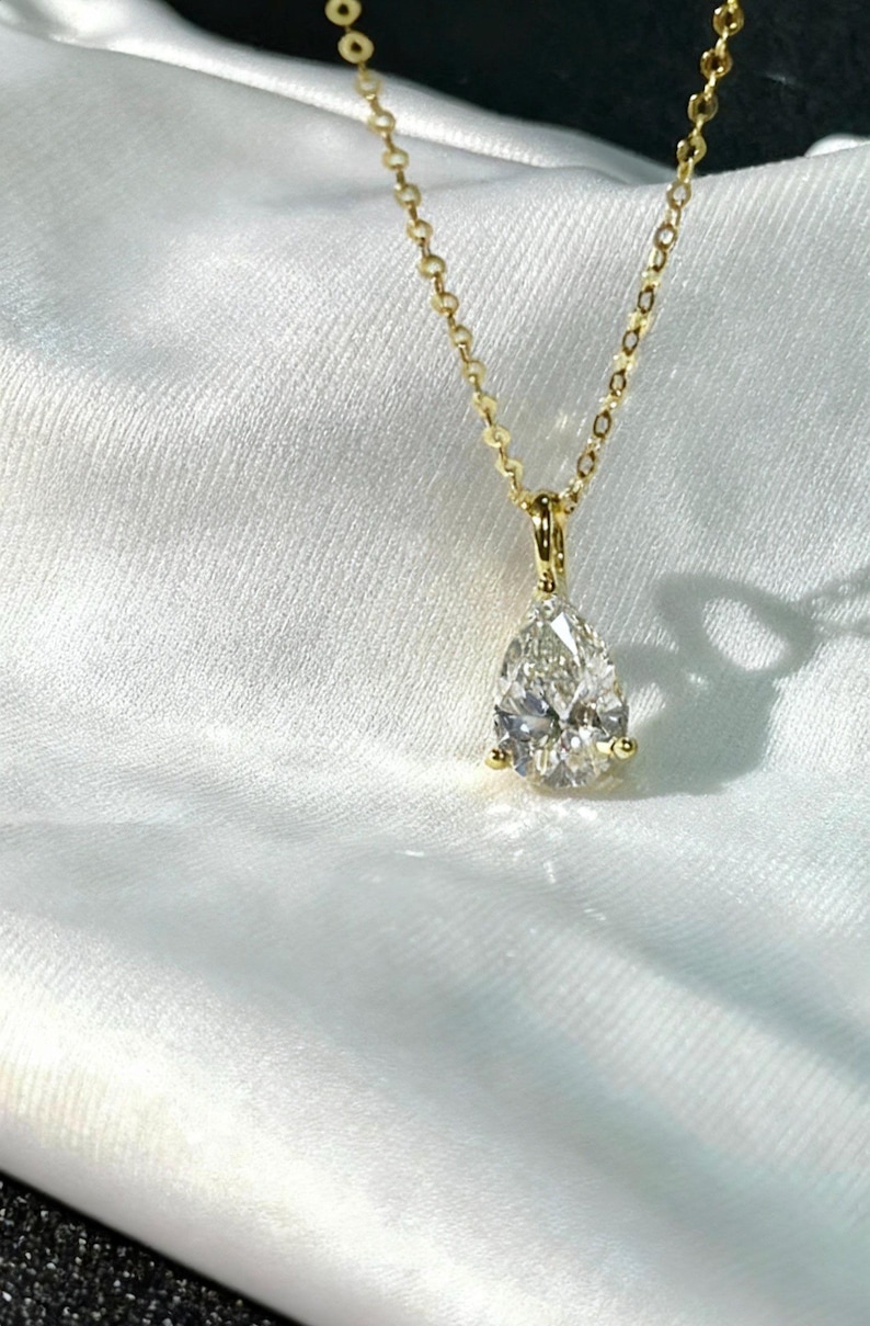 May include: A gold necklace with a pear-shaped diamond pendant. The diamond is clear and faceted, reflecting light. The chain is delicate and gold, with a simple clasp. The necklace is displayed on a white, textured surface.