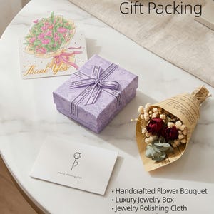 May include: A gift set on a white marble table includes a lavender jewelry box with a ribbon, a card with a flower illustration, a small bouquet, and a jewelry polishing cloth. The words "Gift Packing" are visible.