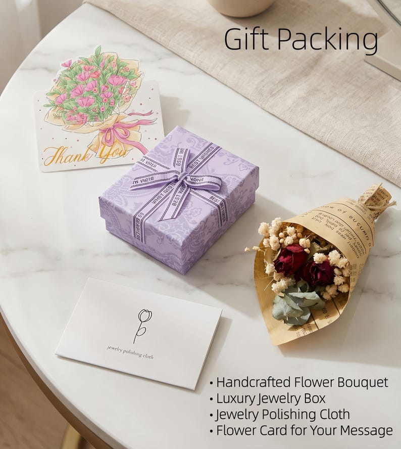 May include: A gift set on a white marble table includes a lavender jewelry box with a ribbon, a card with a flower illustration, a small bouquet, and a jewelry polishing cloth. The words "Gift Packing" are visible.