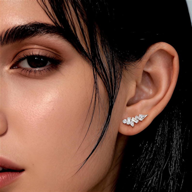 May include: A close-up of a person's ear adorned with a delicate, asymmetrical earring. The earring features multiple clear, sparkling gemstones in various shapes, including round and baguette cuts, arranged in a cluster design.