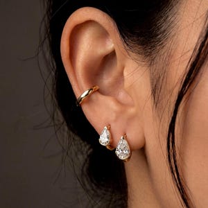 May include: Close-up of a person's ear adorned with gold jewelry. A gold ear cuff is at the top of the ear, and two gold hoop earrings with pear-shaped, clear gemstones are in the earlobes. The jewelry is set against a dark background.