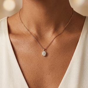 May include: A gold-coloured necklace with an oval pendant set with small, sparkling stones. The necklace is worn against a white garment, creating a contrast that highlights the jewellery.
