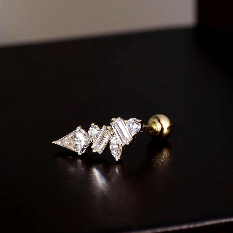 May include: A gold-colored earring with a cluster of clear, sparkling gemstones. The design features a mix of shapes, including a triangle, rectangles, and round stones, creating a delicate, elegant look. The earring has a small, round backing.
