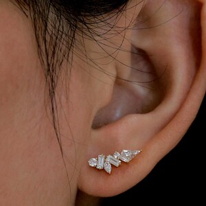 May include: A gold-toned ear climber earring with multiple clear, sparkling gemstones. The gemstones are various shapes, including round, baguette, and pear-shaped, arranged in a cluster design. The earring is worn on the ear.