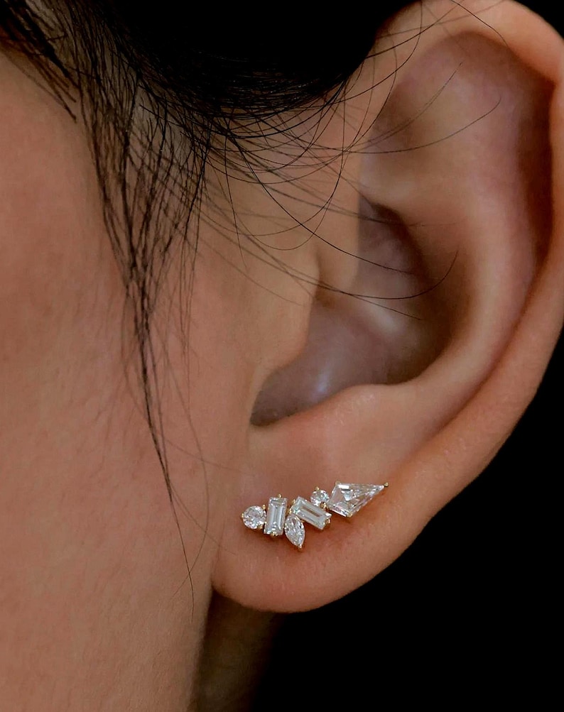 May include: A gold-toned ear climber earring with multiple clear, sparkling gemstones. The gemstones are various shapes, including round, baguette, and pear-shaped, arranged in a cluster design. The earring is worn on the ear.