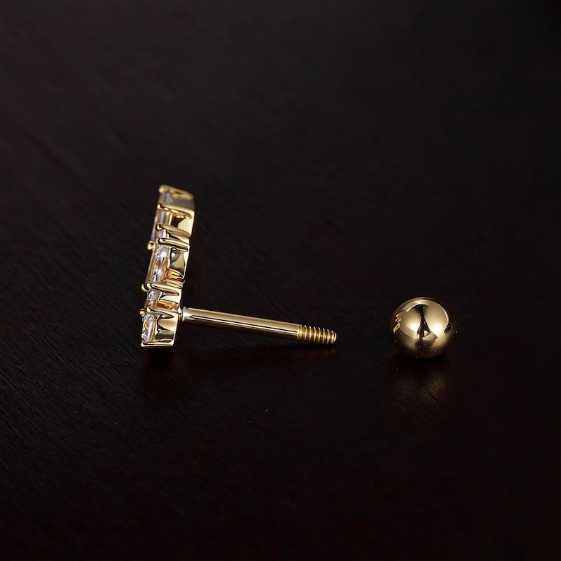 May include: A gold-colored earring with a cluster of clear, rectangular and round gemstones. The earring has a threaded post and a small, round ball backing. The jewelry is set against a dark brown background.