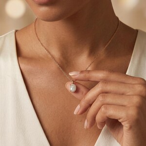 May include: A rose gold necklace with an oval pendant set with small, sparkling stones. The delicate chain is held by a hand. The pendant is a cluster of stones.